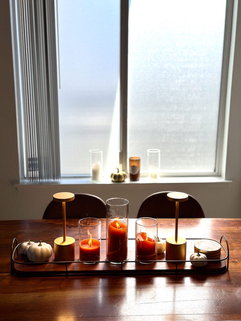 Minimalist fall dining table centerpiece with orange candles, gold LED lamps, white pumpkins, and a wood tray placed in front of a sunlit window.