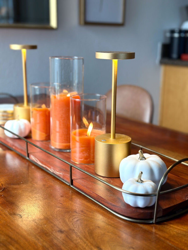 Minimalist fall centerpiece with orange candles, gold LED lamps, and white pumpkins on a wood and metal tray atop a warm wood dining table.