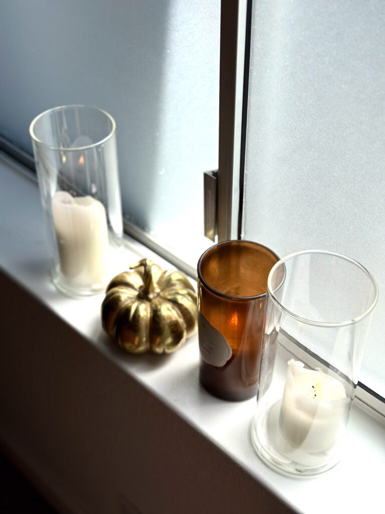 Minimalist fall windowsill with white pillar candles in glass holders, a gold metallic pumpkin, and an amber candle in a brown jar.