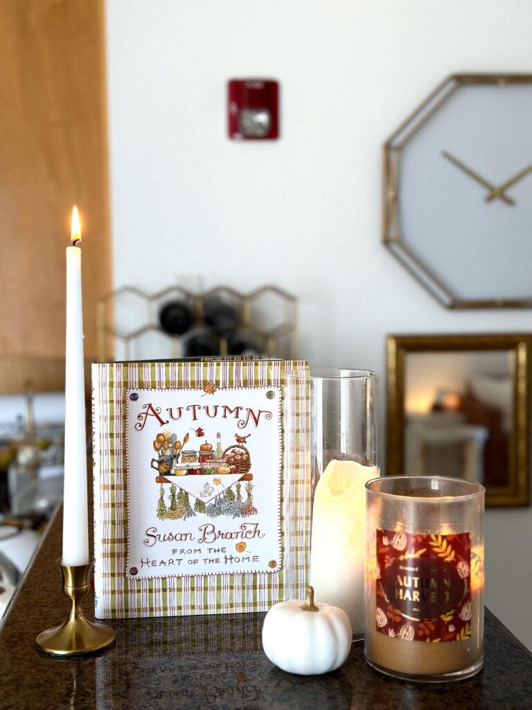 Fall kitchen decor featuring the Autumn cookbook by Susan Branch, white taper candle, seasonal candles, and a mini white pumpkin on a granite countertop.
