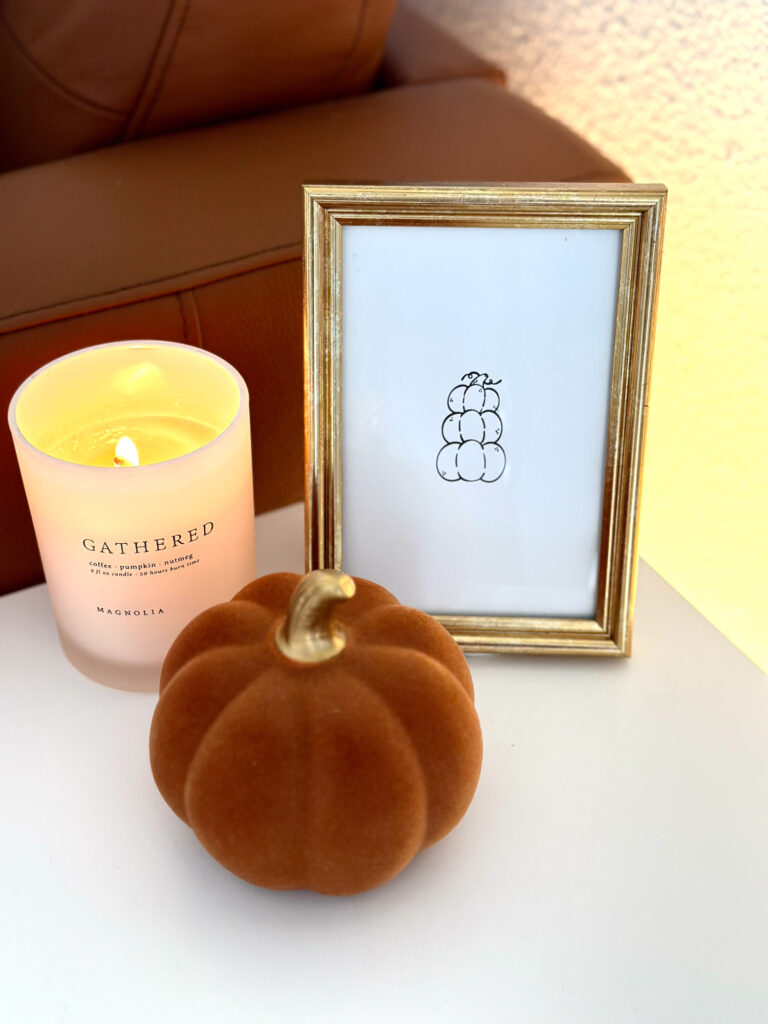 Cozy fall tabletop with a velvet burnt orange pumpkin, lit Gathered candle, and a gold-framed minimalist pumpkin line drawing.