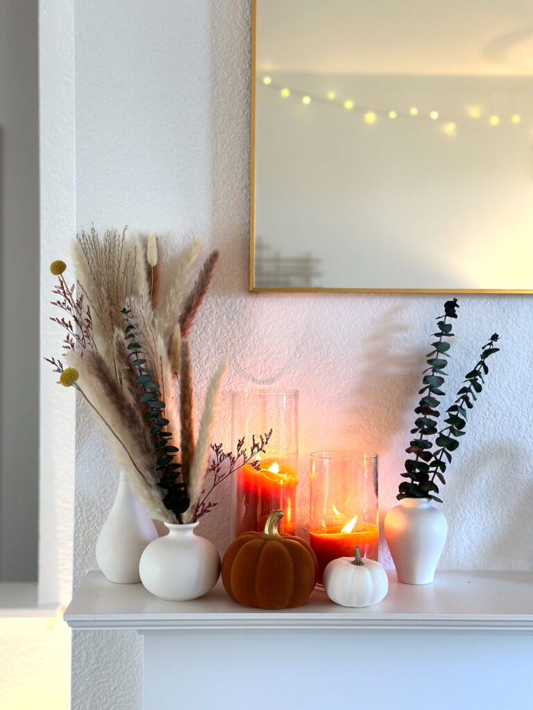 Minimalist fall mantel styled with white ceramic vases, dried pampas grass and eucalyptus, orange candles in glass holders, and velvet pumpkins.