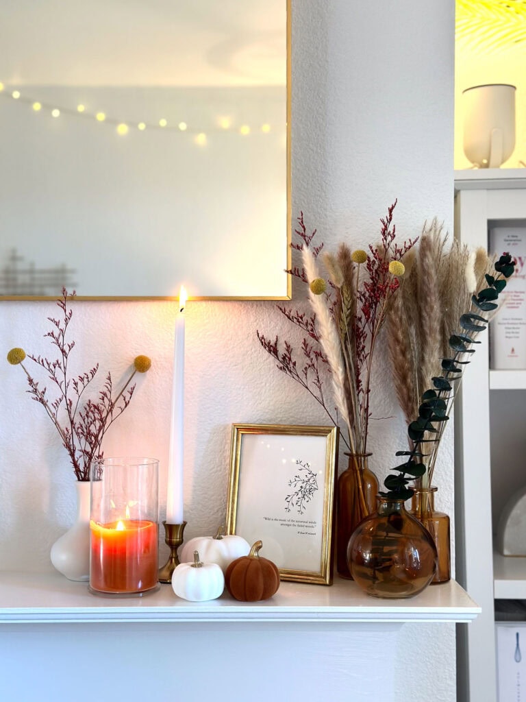 Neutral fall mantel display with amber bud vases, pampas grass, dried florals, white and burnt orange mini pumpkins, lit candles, and framed minimalist artwork.