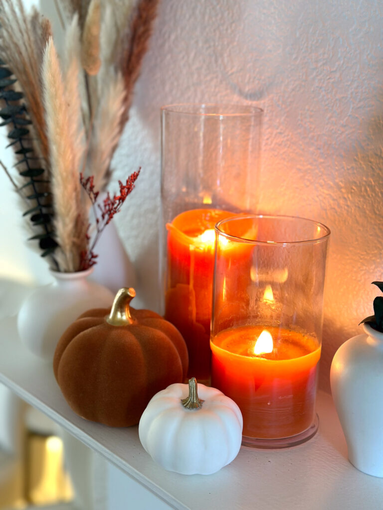 Neutral fall mantel featuring white vases with pampas grass, orange candles in glass holders, and mini pumpkins.