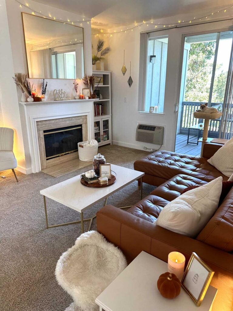 Wide view of a cozy fall living room with a caramel leather sectional, fall-decorated fireplace, coffee table tray, and soft ambient lighting.