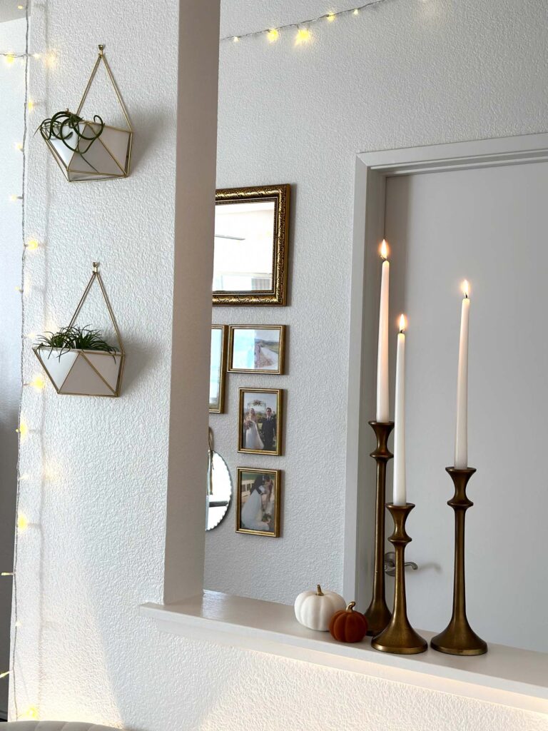 Gold candlesticks with tall white taper candles and two mini pumpkins on a white ledge, with hanging planters, framed photos, and string lights creating a cozy fall atmosphere.