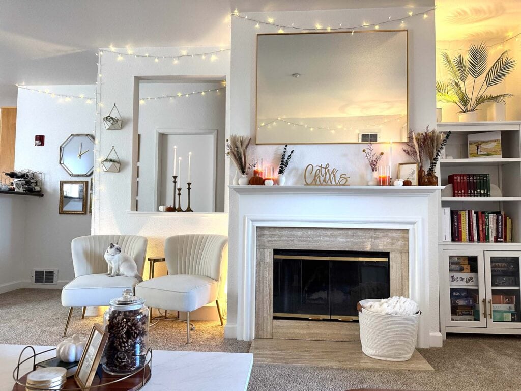 Neutral minimalist fall living room with a styled fireplace mantel, dried florals, candles, and string lights, featuring cozy seating and a white cat on an armchair.