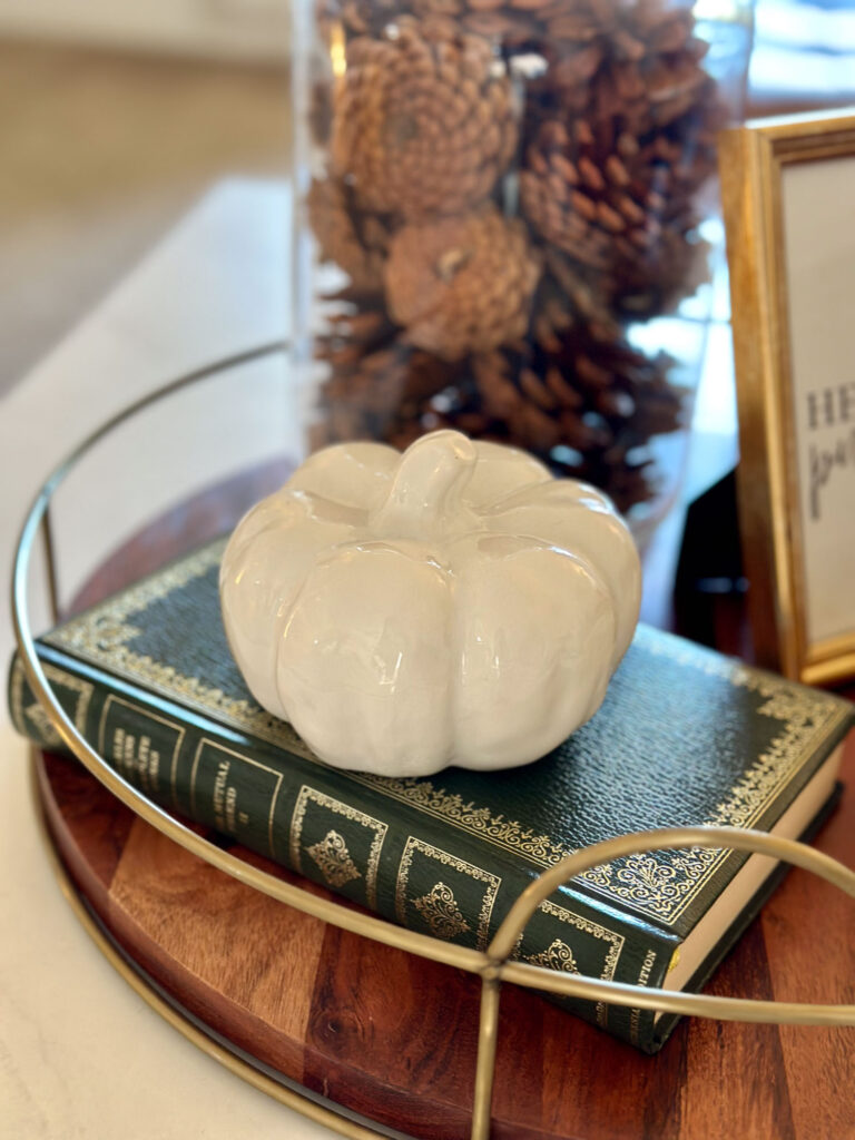 Close-up of a white ceramic pumpkin on a vintage green book, styled on a round fall tray with pinecones in the background.