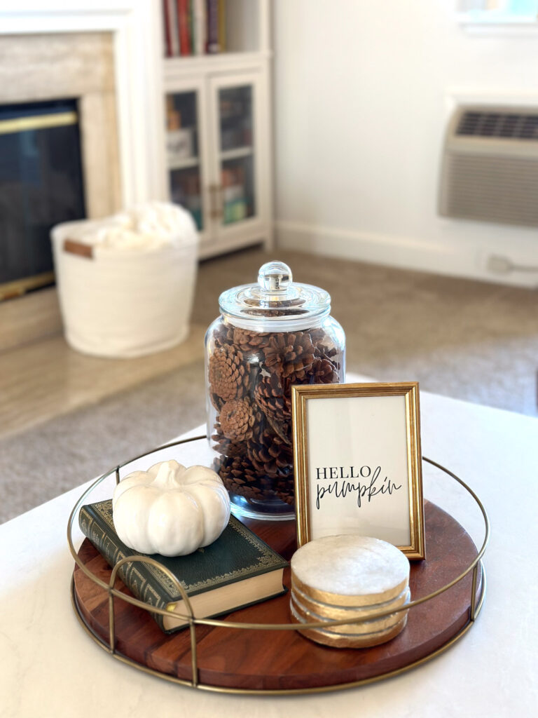 Minimalist fall coffee table tray with a white pumpkin, vintage books, coasters, framed “Hello Pumpkin” print, and pinecones in a glass jar.