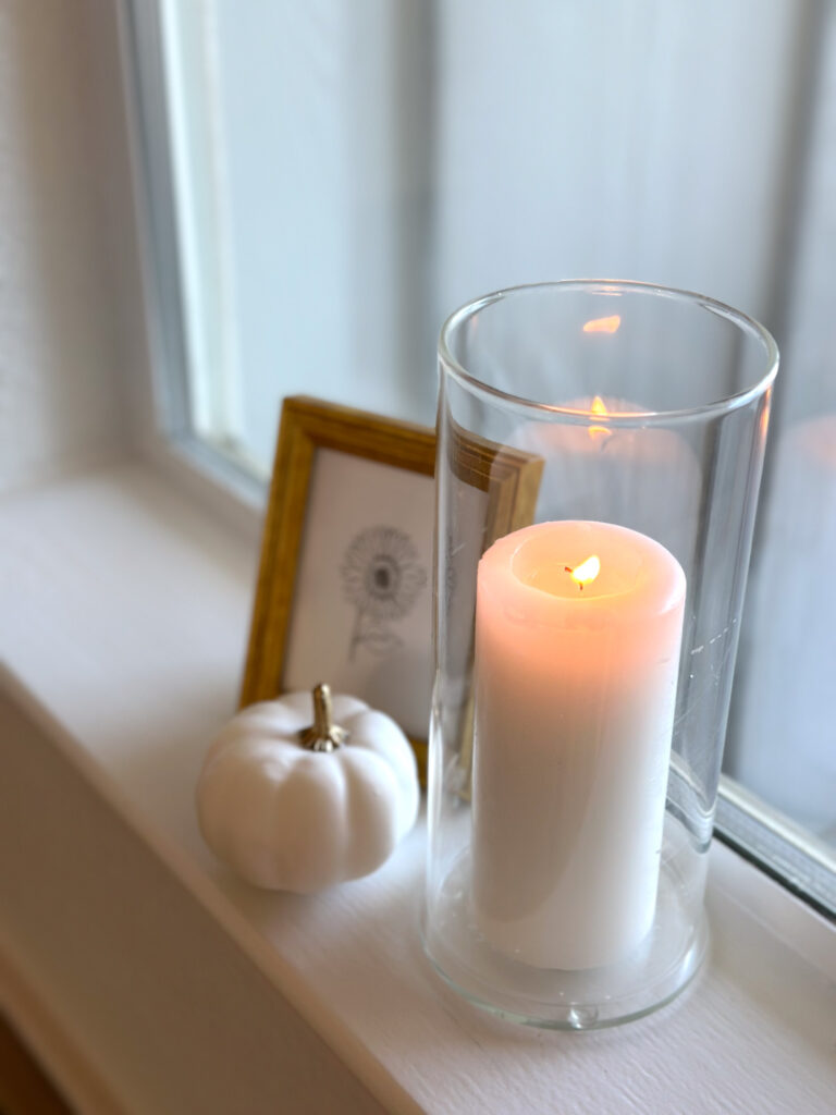 Minimalist fall windowsill with a white pumpkin, pillar candle in a glass holder, and small gold-framed line art print.