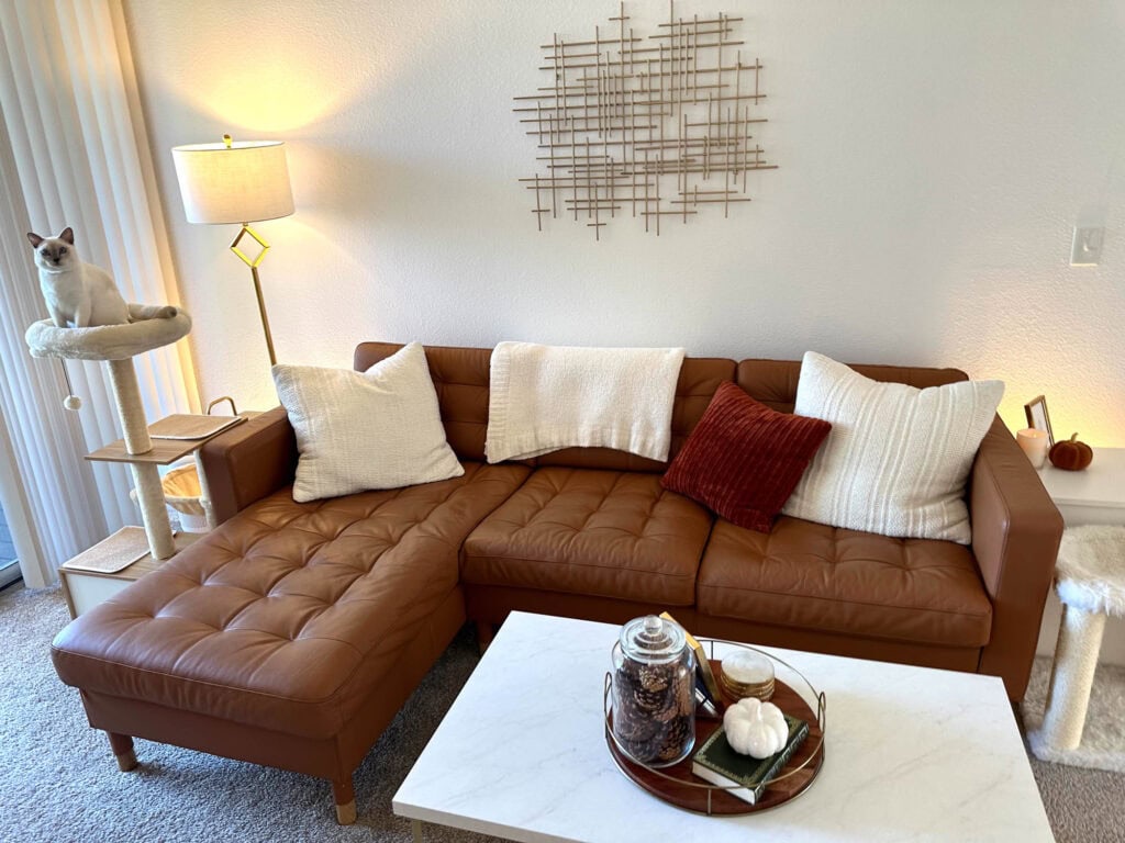 Cozy fall living room with a caramel leather sectional, neutral pillows, burnt orange accent pillow, and a fall tray centerpiece on a marble coffee table. Cat sitting on a perch by the window.