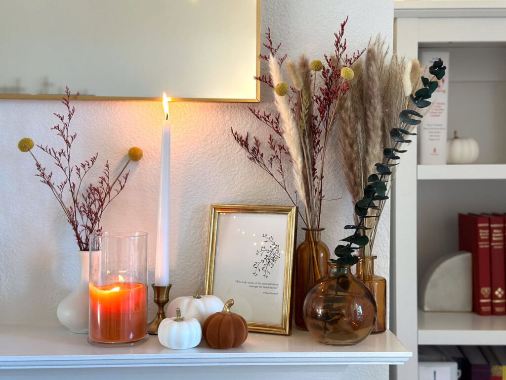 Close-up of a minimalist fall mantel display with amber vases, pampas grass, eucalyptus, white and burnt orange mini pumpkins, candles, and a framed quote.