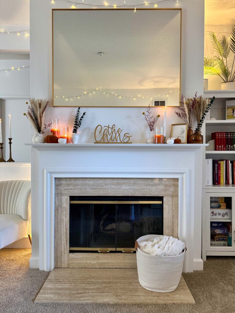 Neutral fall fireplace with pampas grass, amber vases, orange candles, and twinkle lights reflected in a gold-framed mirror.