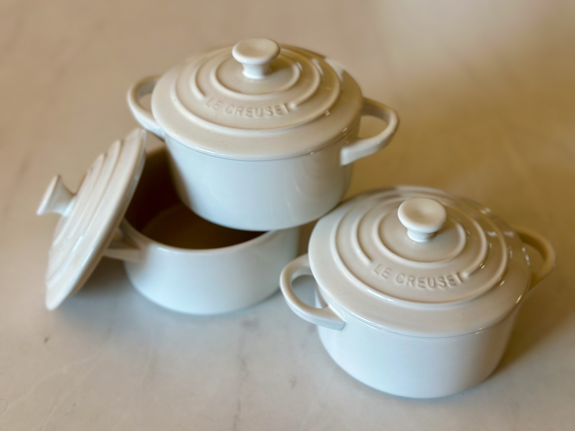 Three white mini Le Creuset cocottes, one with the lid off, stacked on a light countertop