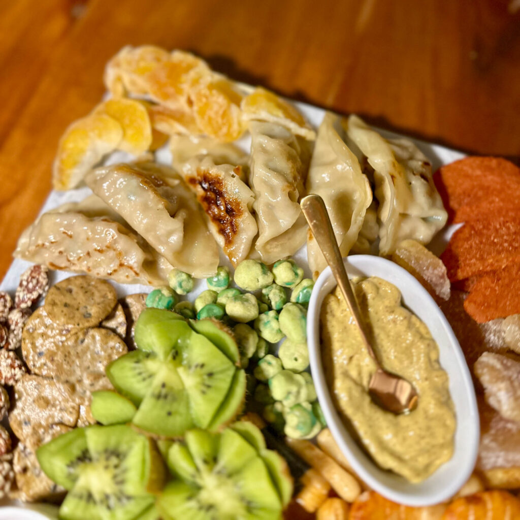 Gyoza dumplings arranged on an Asian charcuterie board alongside kiwi slices, wasabi peas, mustard dip, and dried mandarins.