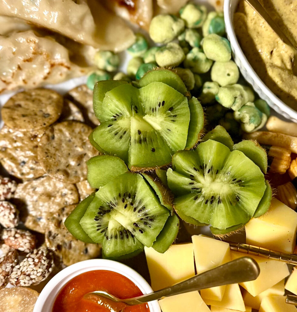 Decorative kiwi slices cut into lotus flower shapes on an Asian charcuterie board, surrounded by cheese cubes, wasabi peas, and crackers.