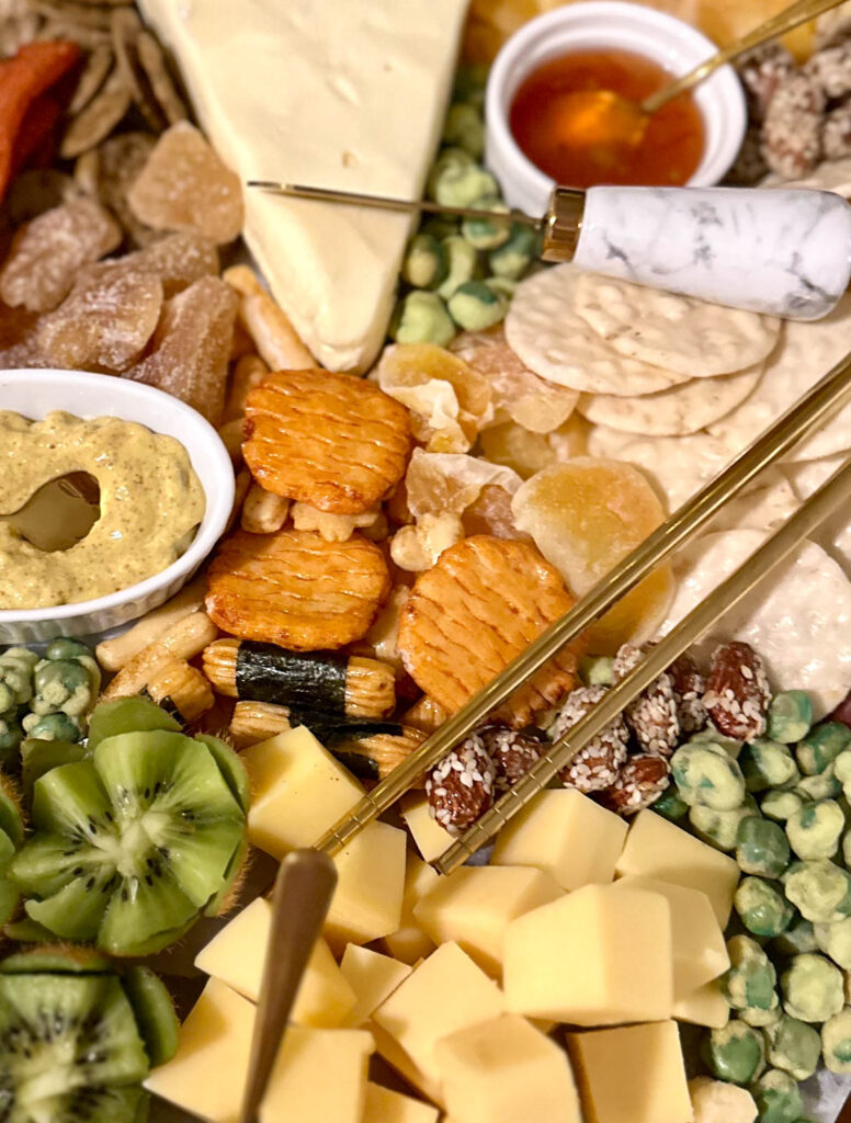 Close-up of the center of an Asian charcuterie board with mustard, rice crackers, crystallized ginger, and sesame snacks.