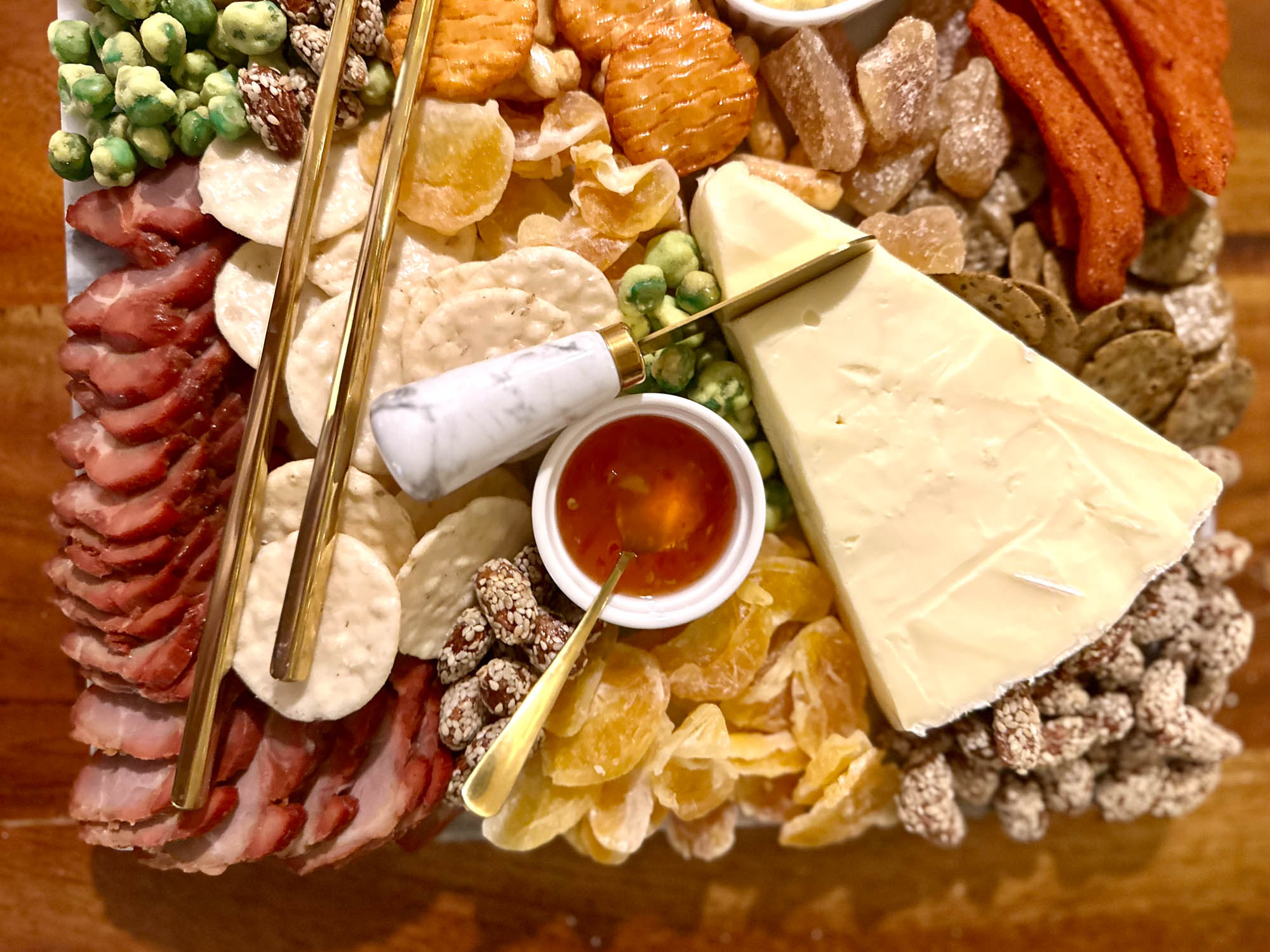 Corner of an Asian charcuterie board with brie, char siu, rice crackers, chili sauce, dried mandarins, and nuts.