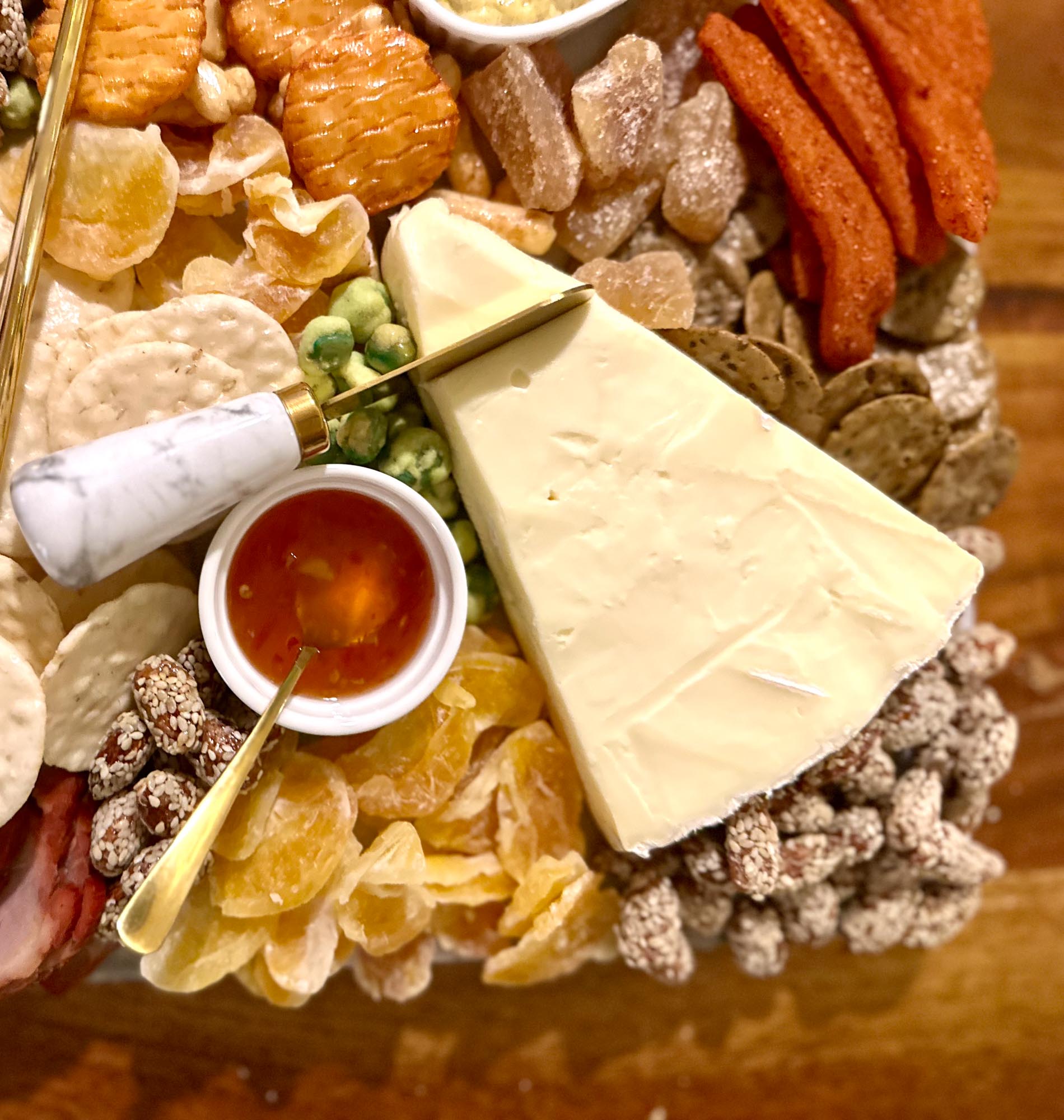 Asian charcuterie board with brie cheese, rice crackers, chili sauce, dried fruit, and spiced nuts arranged on a wooden board.