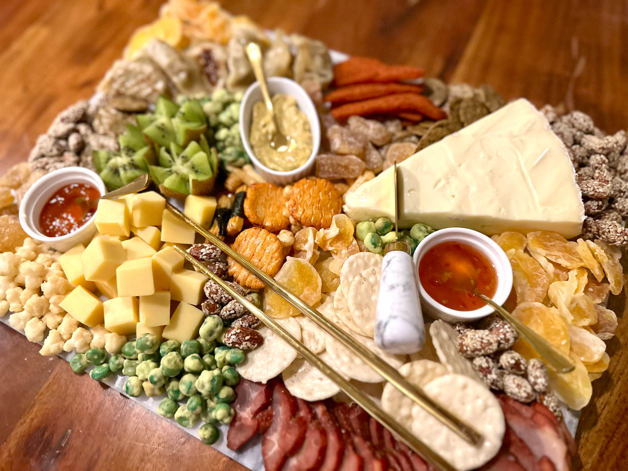 A colorful Asian charcuterie board loaded with cheeses, kiwi, dried fruit, wasabi peas, char siu, and dipping sauces, served with gold chopsticks.