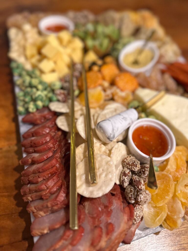 Overhead view of an Asian charcuterie board with sliced char siu pork, brie, rice crackers, dried mandarins, kiwi, nuts, and dipping sauces.