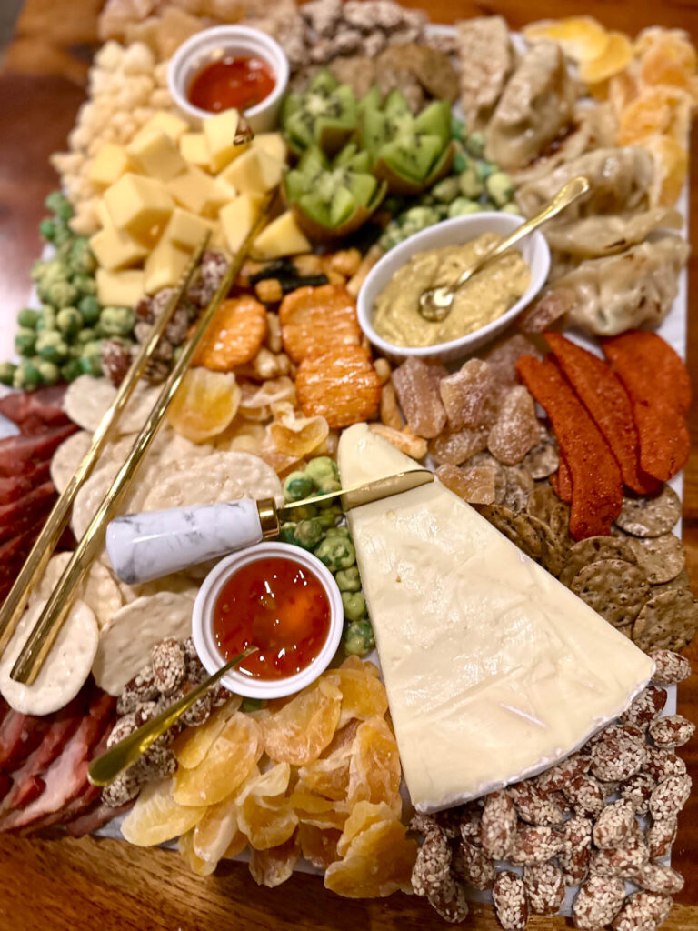 Close up of Asian charcuterie board featuring brie with chili sauce, potstickers and brown mustard, and assorted fruits, cheeses, and nuts.