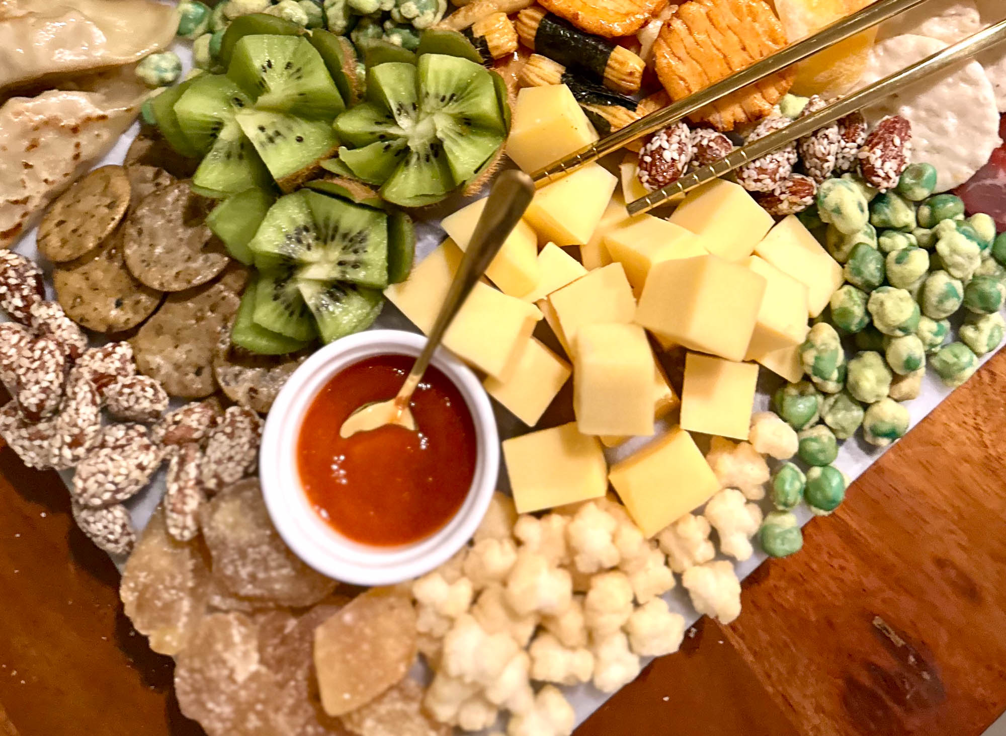 Close-up of an Asian charcuterie board featuring cubed cheddar, sliced kiwi, rice crackers, wasabi peas, and a sriracha dipping sauce.