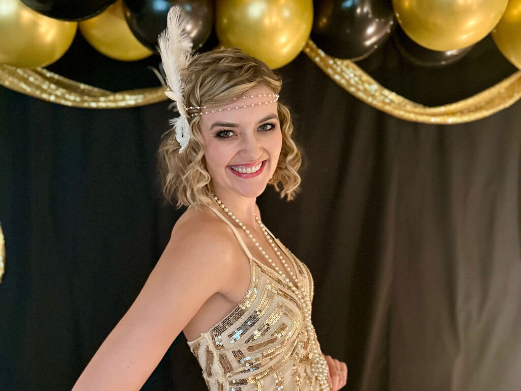 Woman dressed in a gold sequin flapper dress with a feather headpiece, standing in front of black and gold balloons.