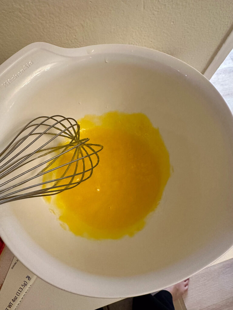 Egg yolks and sugar in a white mixing bowl with a whisk, showing the mixture at the start of whisking for white chocolate crème brûlée.