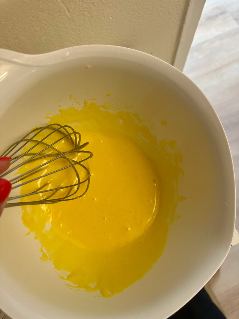 Lightened, smooth mixture of egg yolks and sugar in a white mixing bowl with a whisk after whisking for white chocolate crème brûlée.