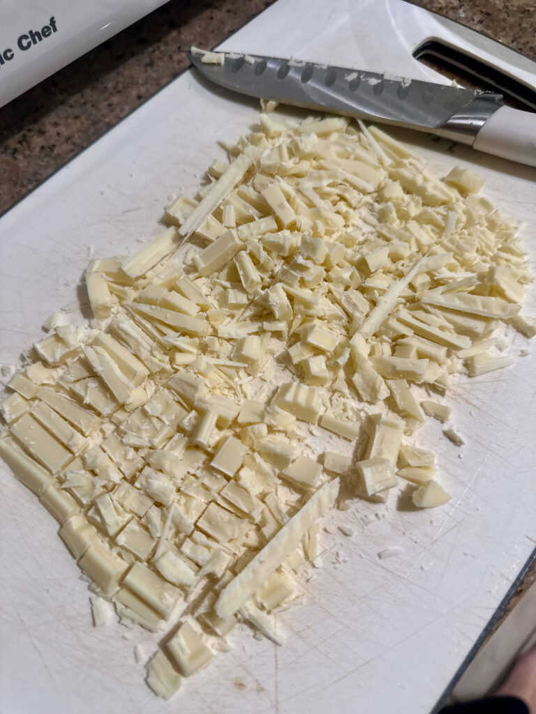 Chopped white chocolate pieces on a cutting board with a chef's knife, ready for melting to make white chocolate crème brûlée