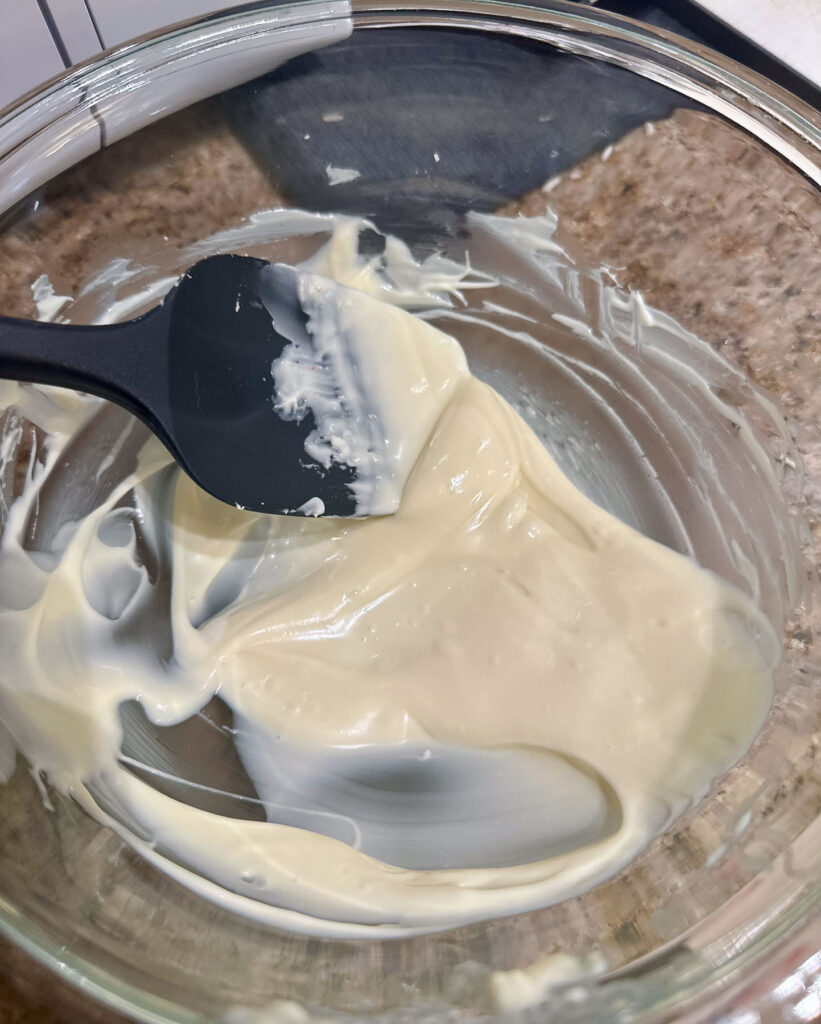 Melted white chocolate in a glass bowl with a black spatula, smooth and glossy after heating for white chocolate crème brûlée