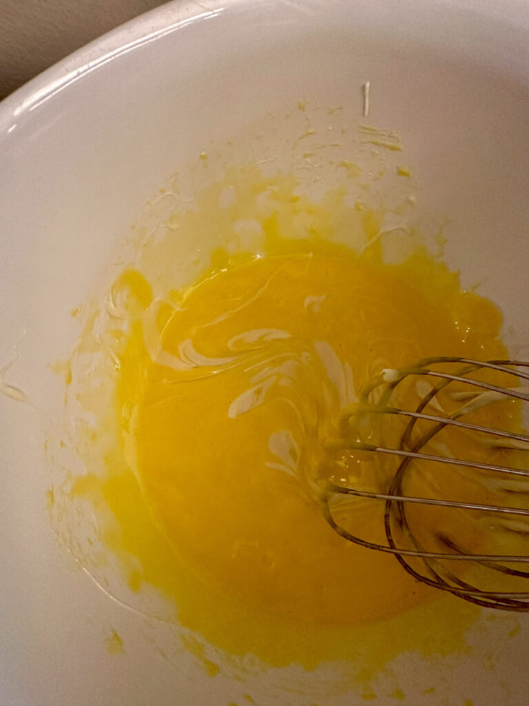 Whisk mixing melted white chocolate into an egg yolk mixture in a white bowl, creating a smooth, pale yellow custard base for white chocolate crème brûlée