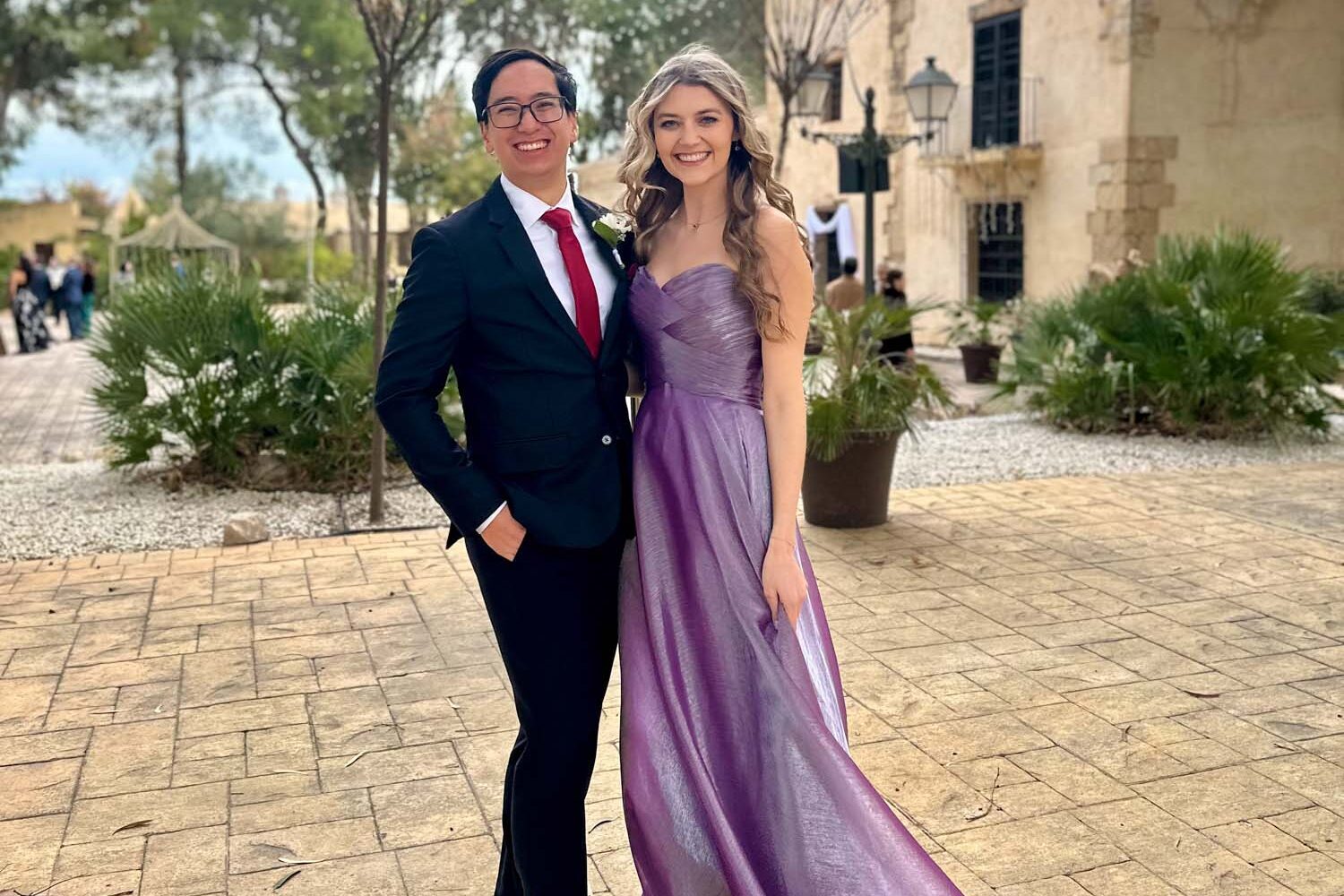 Couple dressed in formal wedding guest attire posing outdoors; woman in a lavender gown and man in a dark suit with red tie.