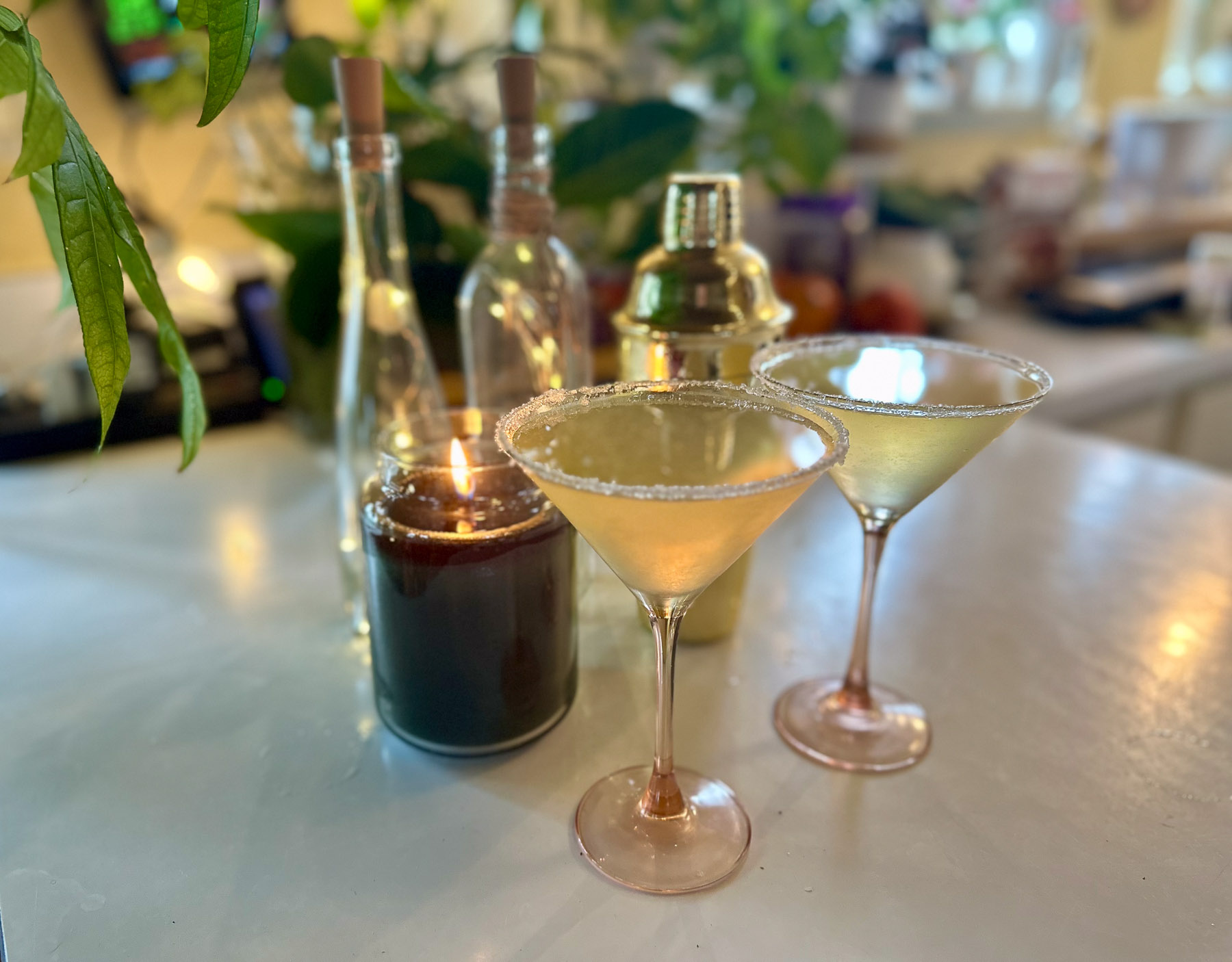 Two pear elderflower martinis with sugared rims on a white countertop, surrounded by candles, glass bottles, and a gold cocktail shaker, with greenery in the background.