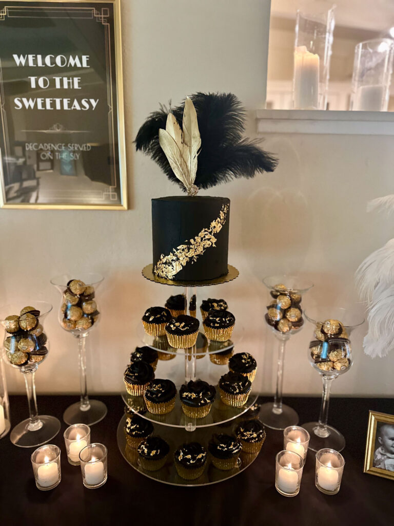 Black and gold “sweet-easy” dessert table with a feathered cake, cupcakes, and candlelight.