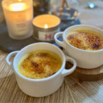 Two small white ramekins of white chocolate cr&egrave;me br&ucirc;l&eacute; with golden, caramelized sugar tops, set on a wooden table beside softly glowing candles in the background.