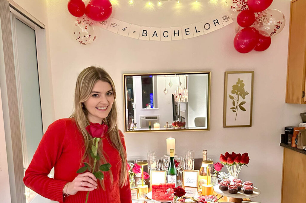 Woman in red holding a rose in front of a Bachelor-themed viewing party setup with balloons, cupcakes, and sparkling wine.