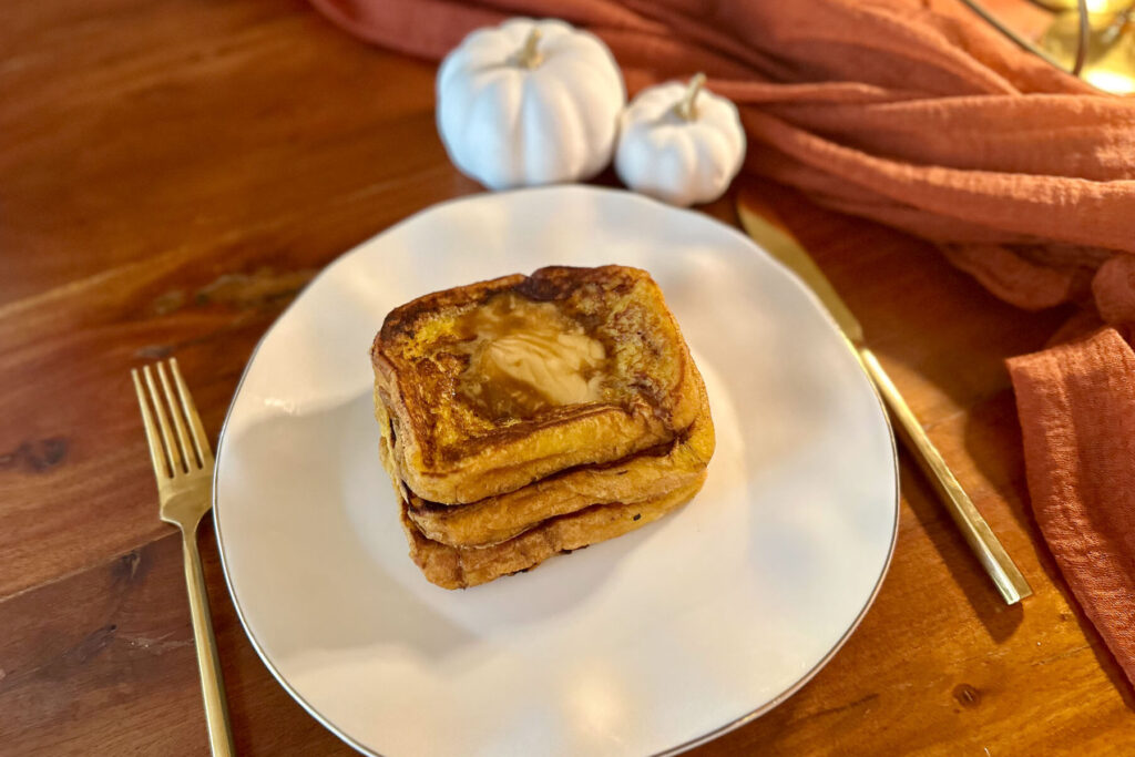 Stack of golden-brown Trader Joe’s pumpkin brioche French toast topped with melting butter on a white plate, with mini white pumpkins and rust-colored cloth in the background.