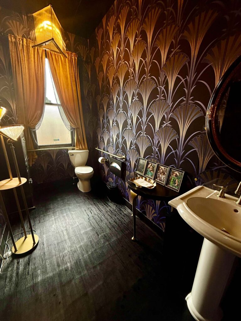 Luxurious bathroom with dark art-deco wallpaper, gold curtains, and vintage furnishings at By the Teashore.
