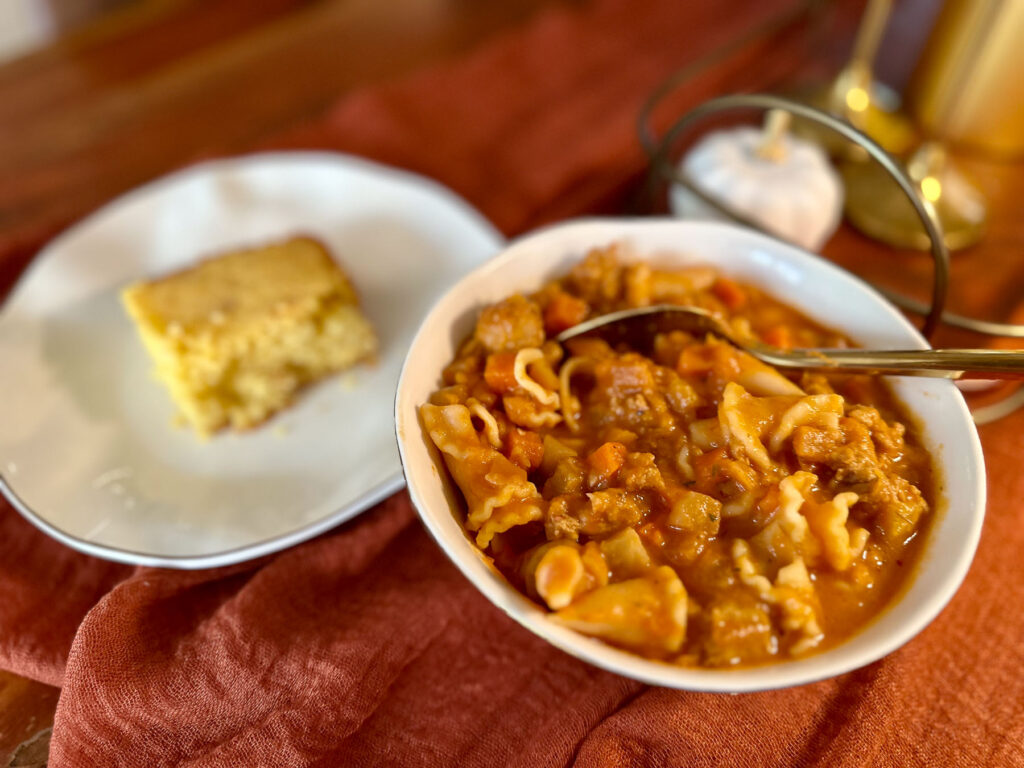 Bowl of hearty Autumnal Harvest soup with pasta and sausage, served alongside a slice of cornbread on a white plate.