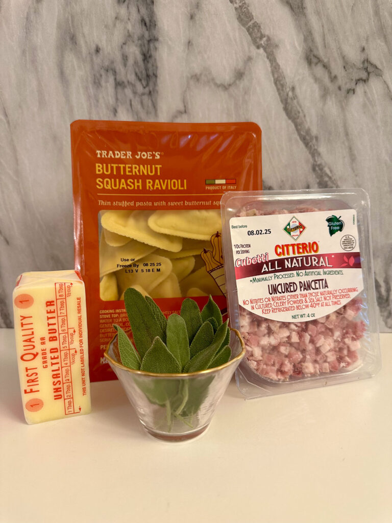 Trader Joe’s butternut squash ravioli, pancetta cubes, fresh sage in a glass bowl, and a stick of butter arranged against a marble backdrop.