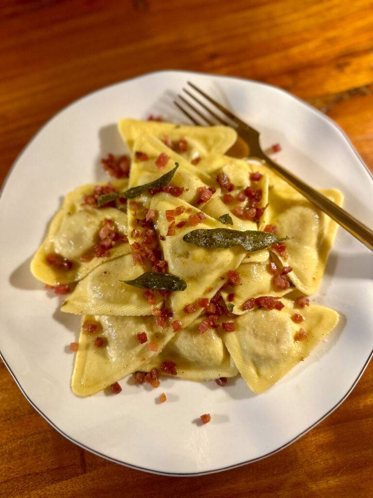 Plate of Trader Joe’s butternut squash ravioli topped with crispy pancetta bits and fried sage leaves, served with a gold fork.