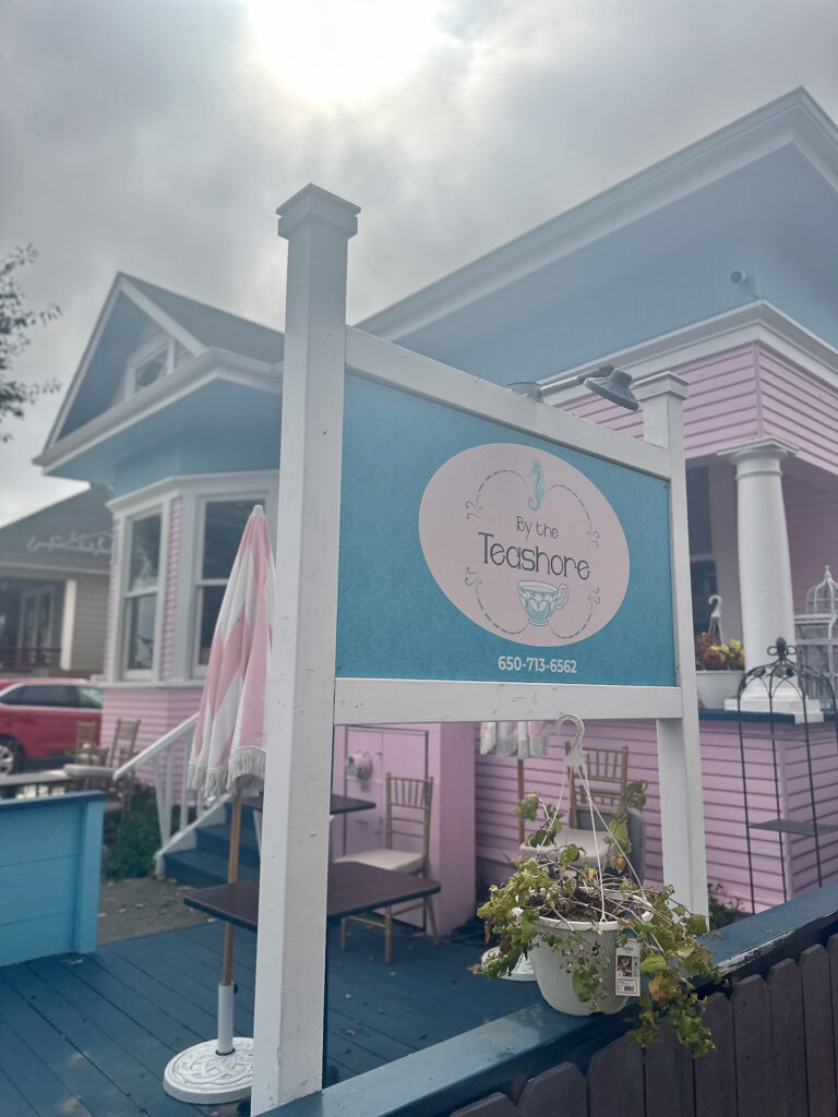 Exterior of By the Teashore in Half Moon Bay, a pink Victorian tea house with teal accents and signage out front.