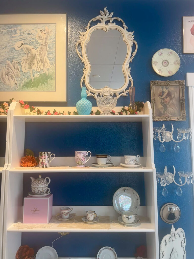 Display shelves with vintage teacups and decor against teal walls at By the Teashore.