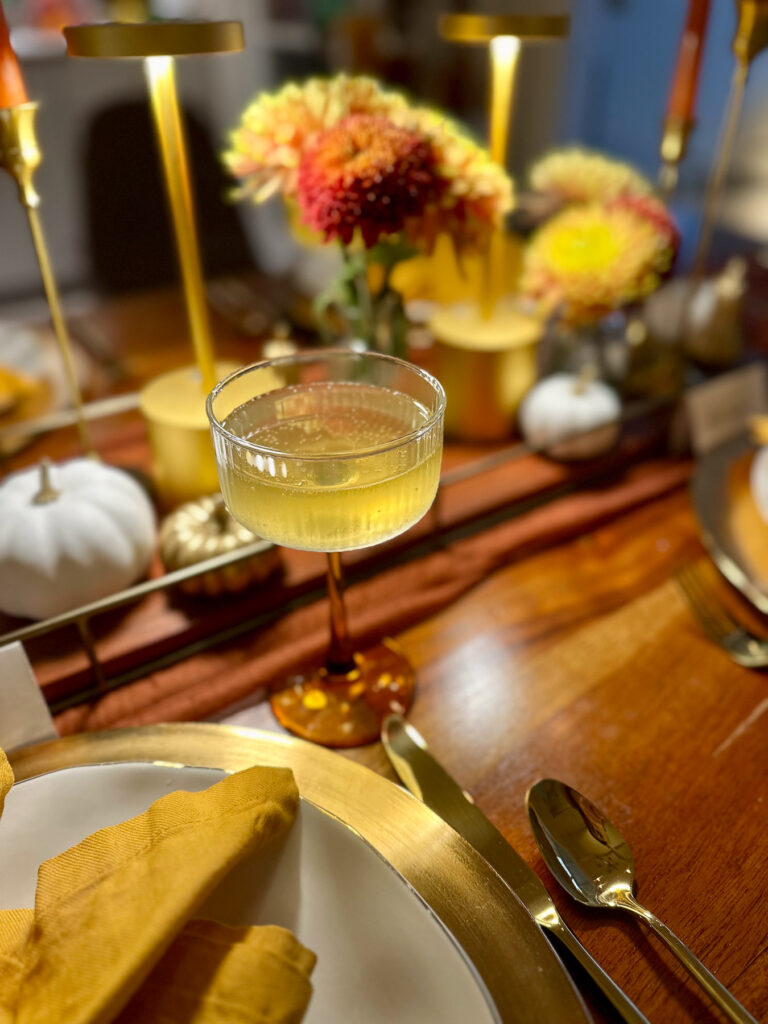 Yellow-stemmed coupe glass cocktail at a fall dinner party table with gold flatware and centerpiece decor.