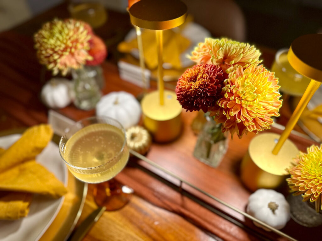 Fall dinner party tablescape with downlights, golden mums, coupe glass, white pumpkins, and candles.