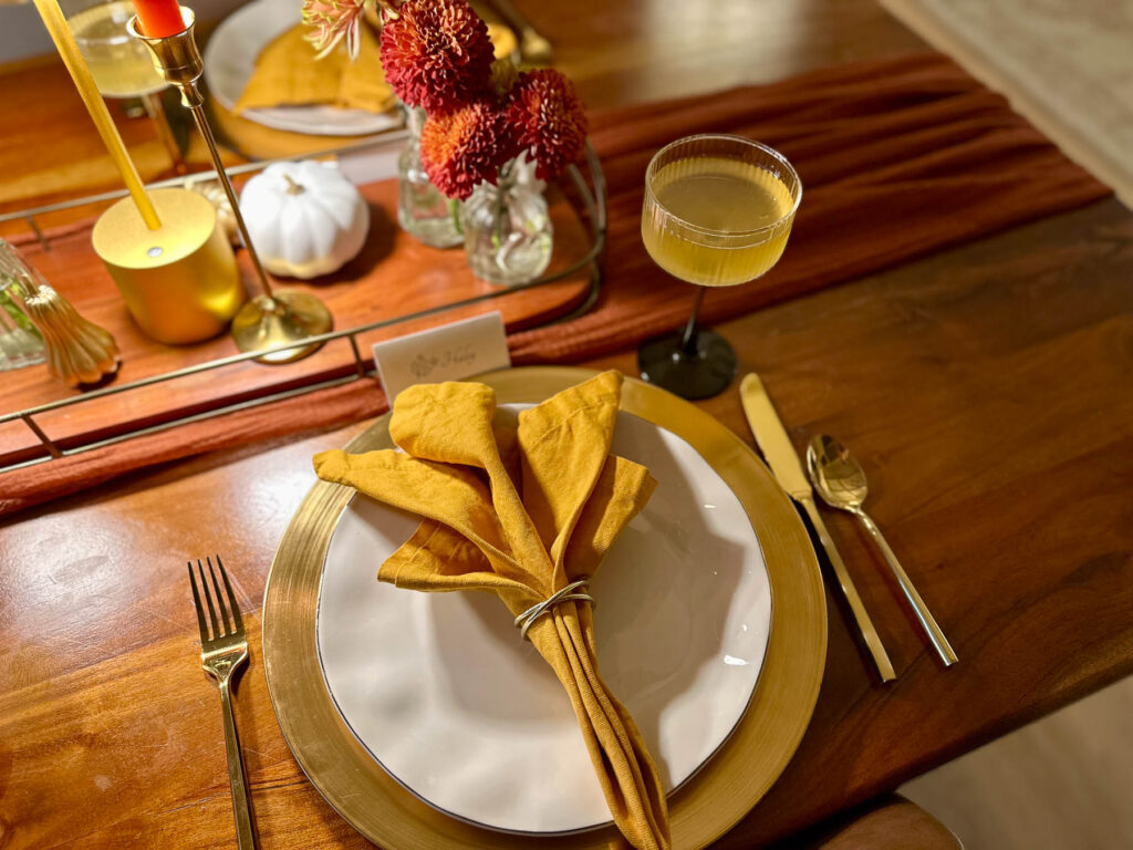 Fall dinner party table setting with gold charger, ochre napkin, cocktail, and autumn centerpiece.