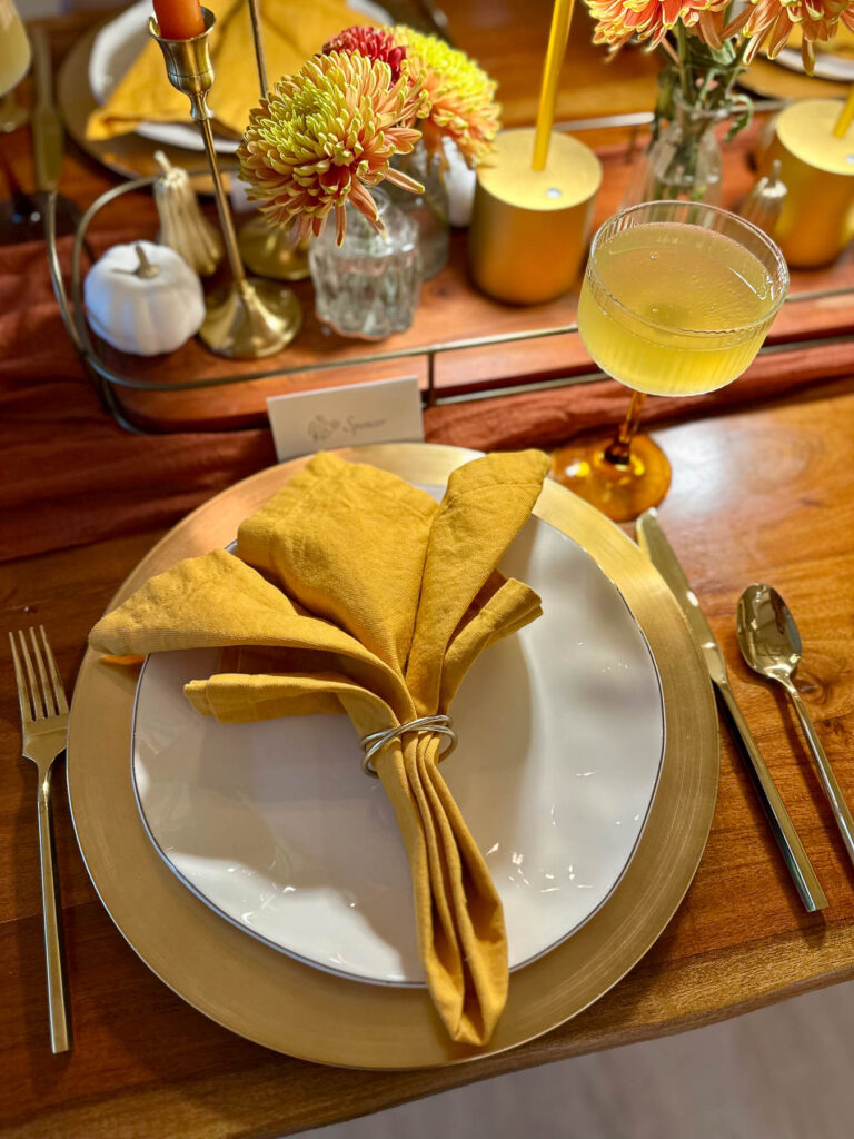 Close-up of a fall place setting with gold charger, ochre napkin, coupe glass, and seasonal centerpiece.
