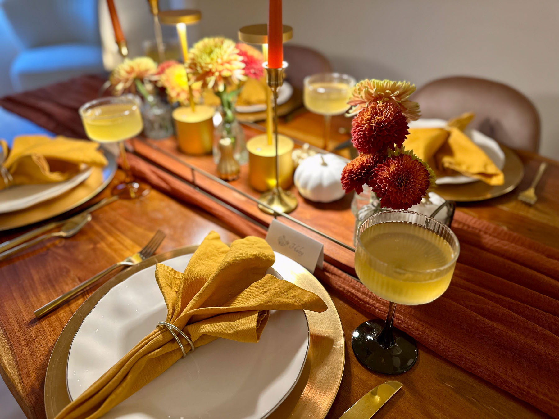 Elegant fall dinner party place setting with gold charger, mustard napkin, coupe glass with cocktail, and autumn floral centerpieces with candles.